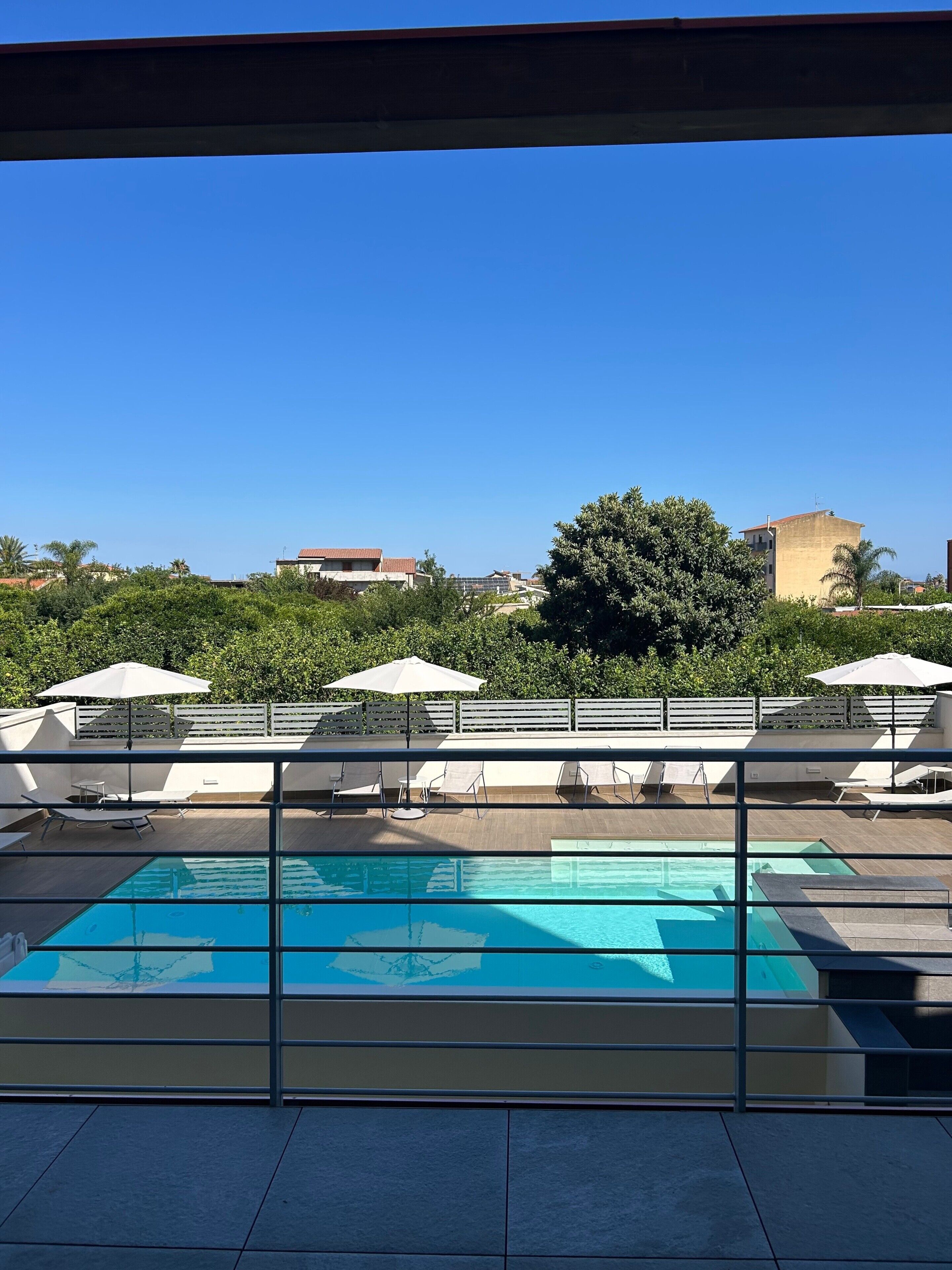 A rooftop pool area with lounge chairs and umbrellas, surrounded by greenery and buildings.