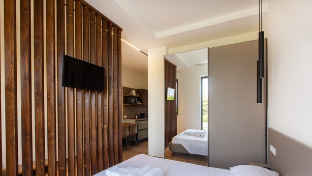 Habitación de hotel con cama, televisor montado en un panel de madera, kitchenette y baño visible a través de una puerta abierta.