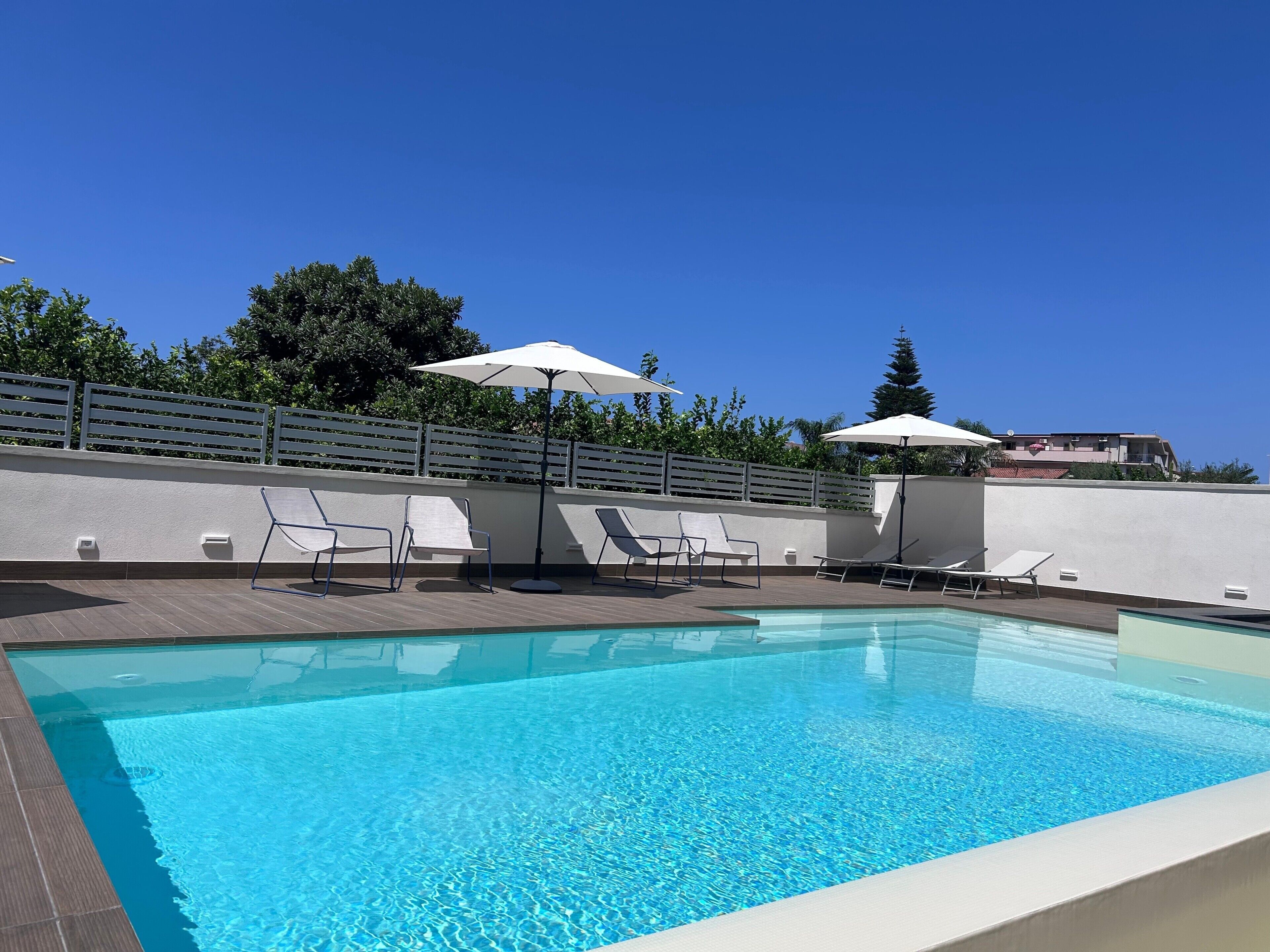 Piscina con sillas de descanso y sombrillas, rodeada por una valla blanca y árboles.