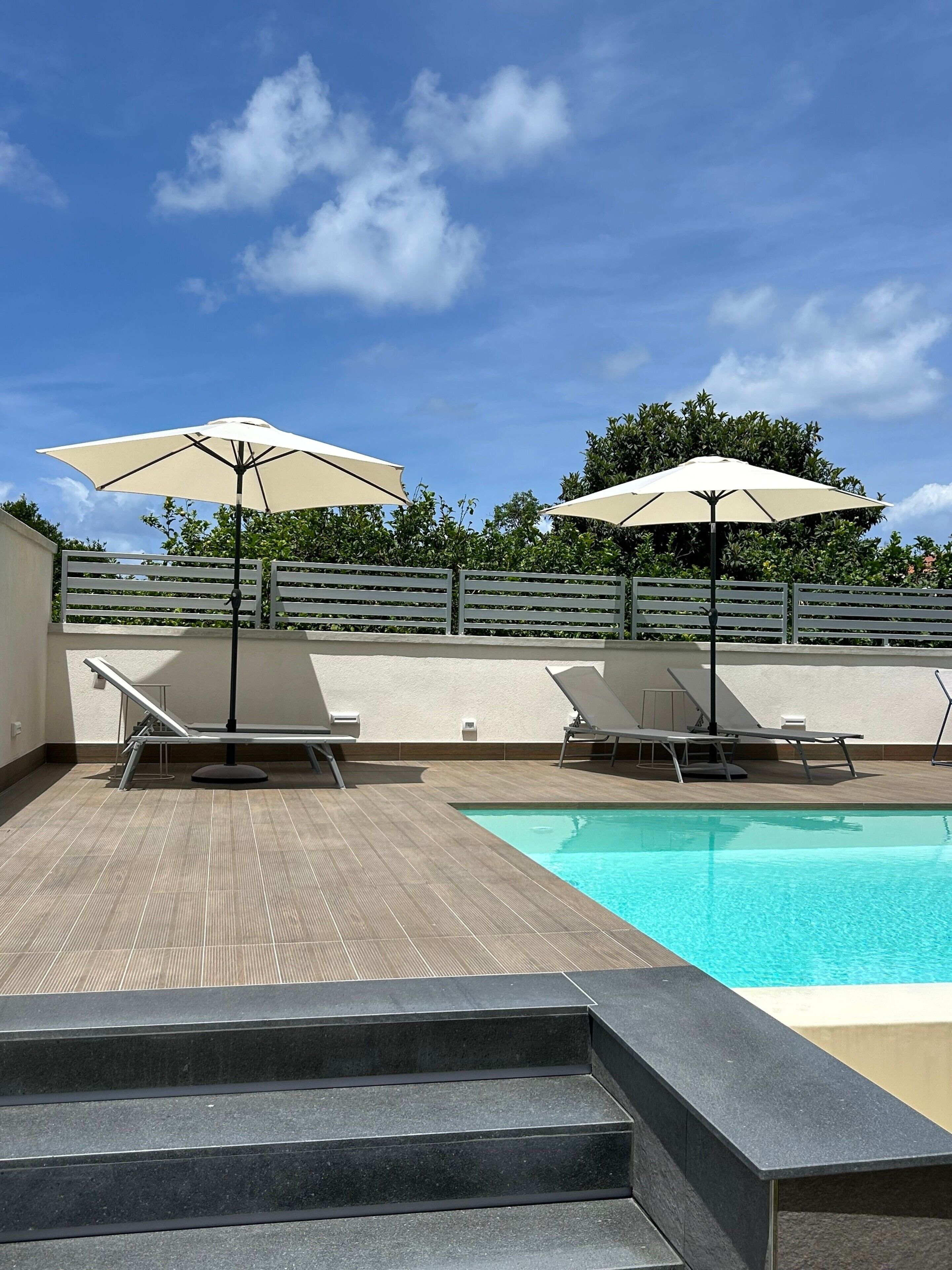 A swimming pool with lounge chairs and umbrellas, surrounded by a wooden deck and a low wall.