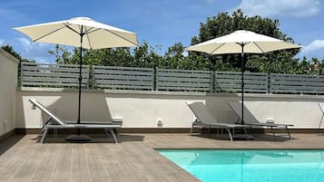 A swimming pool with lounge chairs and umbrellas, surrounded by a wooden deck and a low wall.