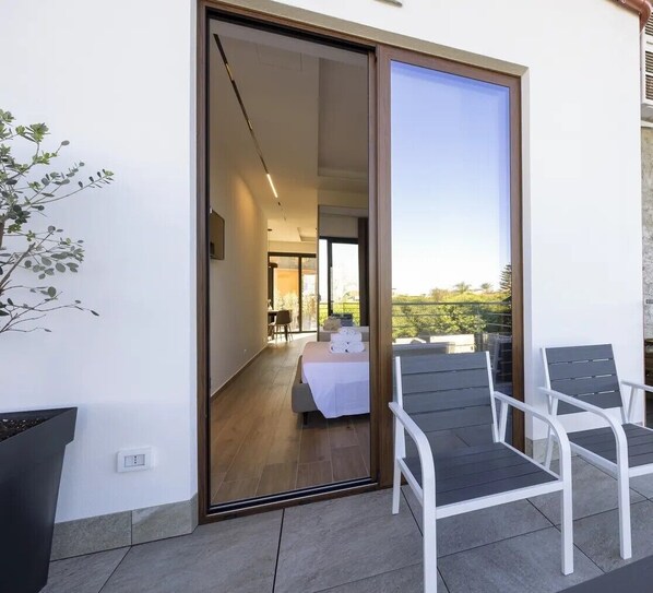 A modern hotel room with a sliding glass door leading to a balcony with two chairs and a bed with a white bedspread.