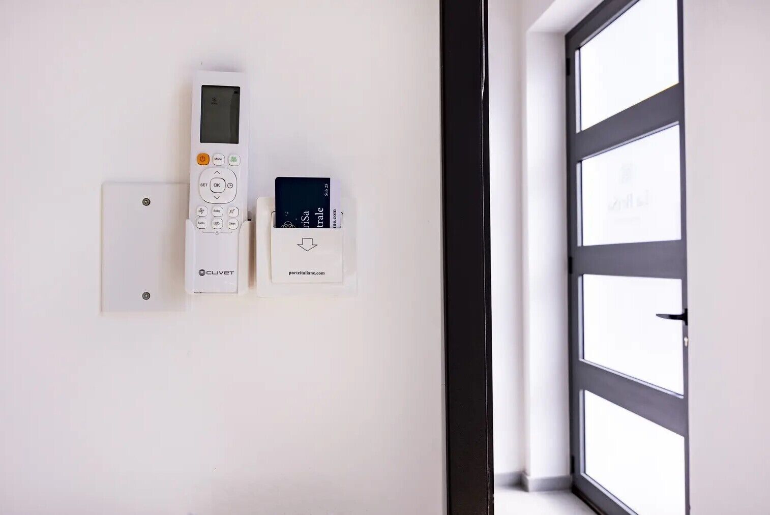 Un panel de control montado en la pared con pantalla digital y botones, una tarjeta con una dirección web, y una puerta con ventana.