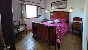 Un dormitorio con una cama de madera, una silla, una mesita de noche, un ventanal y un cuadro en la pared.