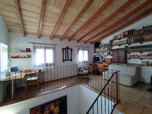 Un vaste espace de vie doté d’un haut plafond, de poutres en bois, d’un escalier, d’un coin repas et d’une bibliothèque remplie de livres.