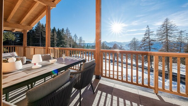 Une terrasse en bois avec une table et des chaises, d’où l’on peut admirer un paysage enneigé parsemé d’arbres et dominé par des montagnes.