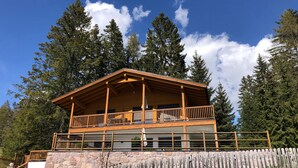 Une cabane en bois dotée d’un balcon, construite sur des fondations en pierre et entourée de grands arbres.