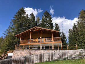 A wooden cabin with a balcony, stone foundation, and surrounded by tall trees.