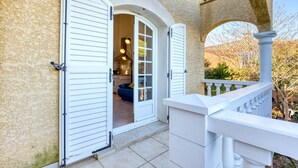 Een huis met een witte gevel, een balkon met een witte balustrade en een deur die leidt naar een kamer met een blauwe bank.