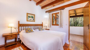 Quarto com cabeceira de madeira, cama com roupa de cama branca, duas mesas de cabeceira com abajures, janela com cortinas e vista para o verde lá fora.
