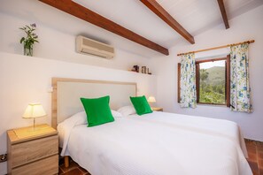 Un dormitorio con cabecera de madera, dos almohadas verdes, una colcha blanca, una mesita de noche con lámpara, una ventana con cortinas florales y vistas al exterior con vegetación.