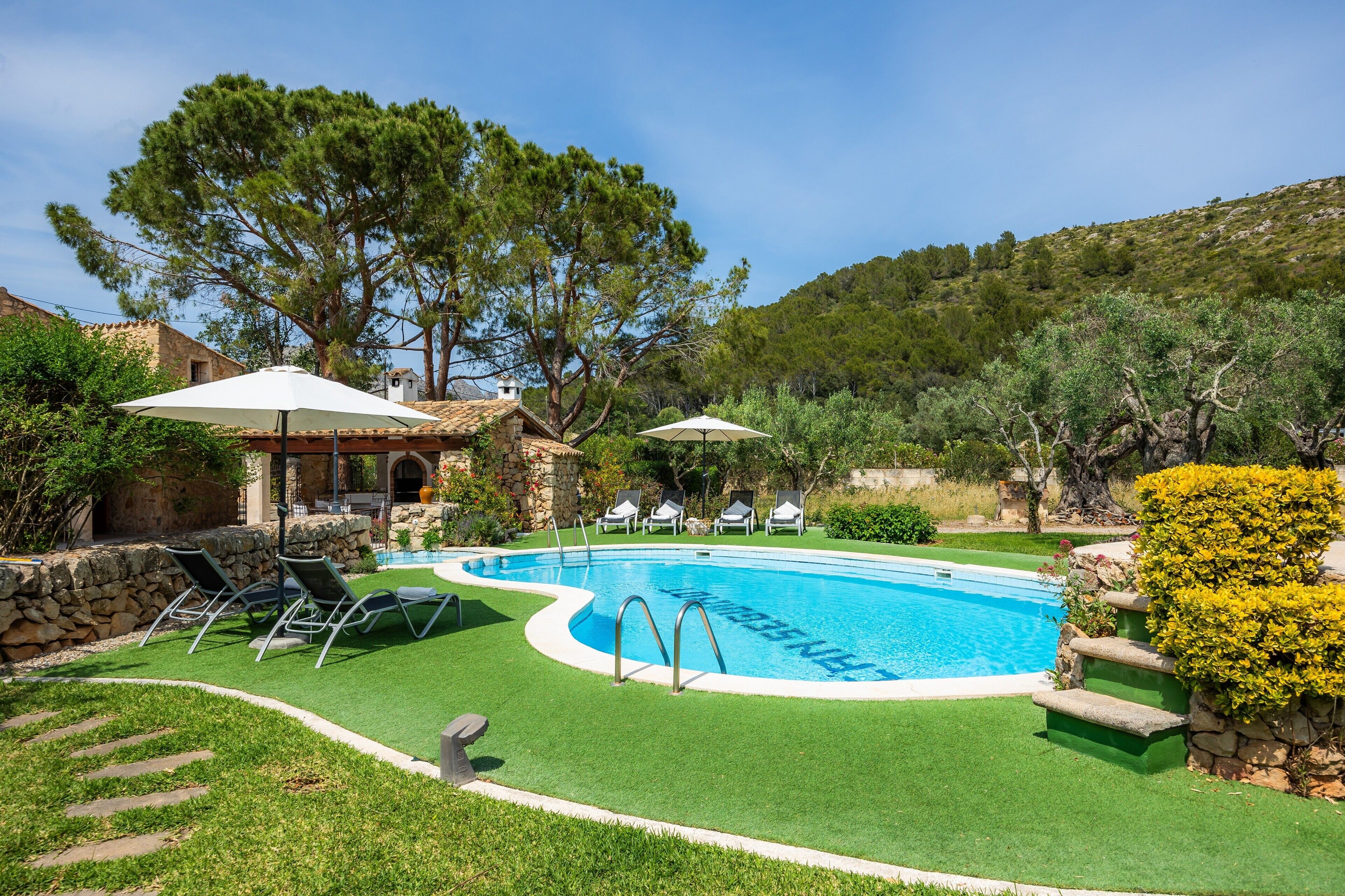 A swimming pool with a stone wall, a lounge area with chairs, and a view of a hill.