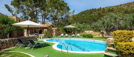 A swimming pool with a stone wall, a lounge area with chairs, and a view of a hill.