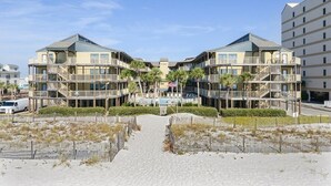 Un hotel frente a la playa con un patio central, varios balcones y un camino de arena que conduce a la playa.