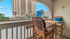 A balcony with a wooden table and chairs, overlooking a beachfront with high-rise buildings and palm trees.