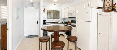 Una cocina con una mesa redonda y cuatro taburetes, muebles blancos, un microondas y un refrigerador.