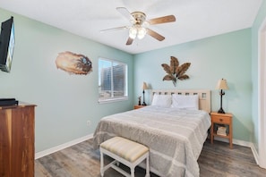 A bedroom with a bed, two bedside tables, a bench, a ceiling fan, a wall-mounted TV, and a window with blinds. - Las Palmas 114- Pool, BBQ, short walk to beach (Gulf Shores)