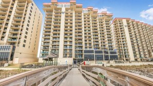 A multi-story residential building with balconies, a central entrance, and a wooden boardwalk leading to the entrance.