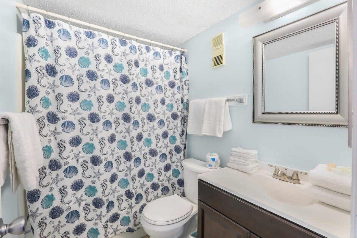 Un baño con cortina de ducha con diseño de conchas marinas, inodoro blanco, lavabo con espejo y un perchero.