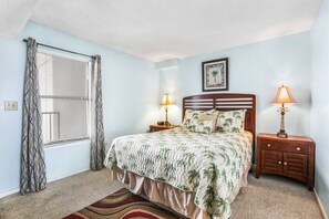 A bedroom with a bed, two bedside lamps, a window with curtains, a framed picture on the wall, and a patterned rug. - Island Wind West 177, Next to Hangout, Beachfront (Gulf Shores)