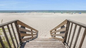 En træpromenade, der fører ud til en sandstrand under en klar himmel.