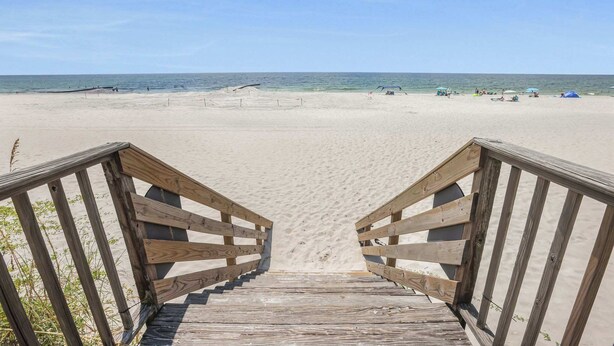 Ein hölzerner Steg führt zu einem Sandstrand unter blauem Himmel.