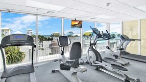 Ein Fitnessstudio mit Laufbändern, einem Crosstrainer und einem stationären Fahrrad, große Fenster mit Blick auf Gebäude und Palmen.