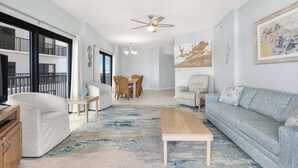 A spacious living room with a large sofa, a coffee table, and a dining area with a table and chairs. There is a ceiling fan and artwork on the walls.