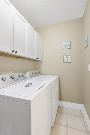 A laundry room with a front-loading washer and dryer, beige walls, and two framed pictures on the wall. - Regency Isle 806 | Gulffront Condo w/Stunning Views, Pools & Sauna (Orange Beach)