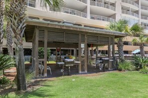 A covered outdoor seating area with tables and chairs, surrounded by palm trees and a building with multiple balconies in the background. - Regency Isle 806 | Gulffront Condo w/Stunning Views, Pools & Sauna (Orange Beach)