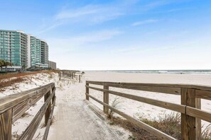 A wooden boardwalk leading to a beach with a high-rise hotel in the background.