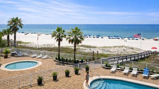 A pool area with lounge chairs, a small pool, and a view of the beach with umbrellas and a flag. - Windward Pointe 305: 3rd floor, Beachfront, pool (Orange Beach)