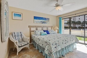 A bedroom with a bed, a chair, a ceiling fan, and a window with a view of buildings and a white fence. - Seaside Beach 2103- Ground level, beachside (Orange Beach)