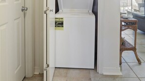 A laundry room with a white washer and dryer, a door with a handle, and a wicker table with chairs. - Seaside Beach 2103- Ground level, beachside (Orange Beach)