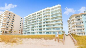 A beachfront hotel with multiple balconies, a sandy beach in the foreground, and a clear sky.