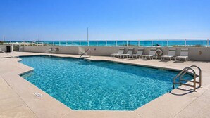 A swimming pool with clear blue water, surrounded by lounge chairs and a lifeguard stand, with a beach view in the background.