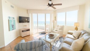 A spacious living room with a large window overlooking the beach, a ceiling fan, and a flat-screen TV mounted on the wall.