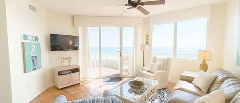 A spacious living room with a large window overlooking the beach, a ceiling fan, and a flat-screen TV mounted on the wall.