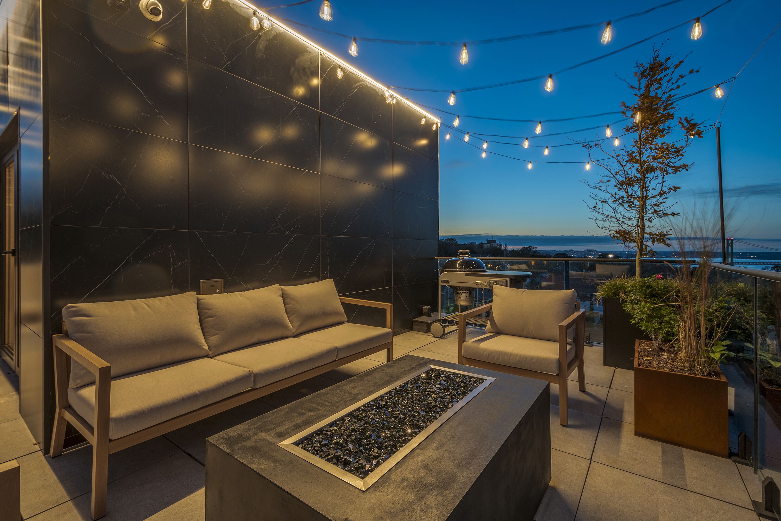 A modern outdoor seating area with a sofa, chairs, and a coffee table, featuring a city view and string lights.