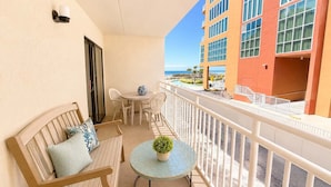 A balcony with a wooden bench, a small table with a potted plant, and a view of a building with large windows and a pool.