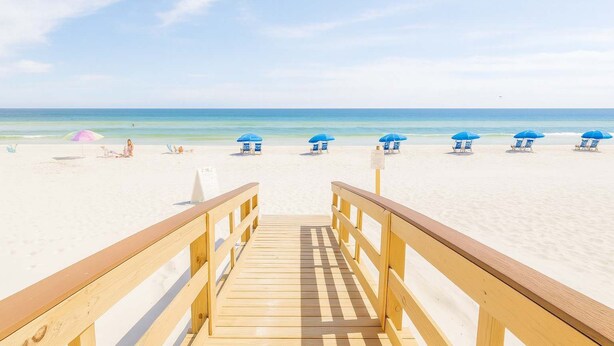 Een houten looppad dat naar een strand leidt, met blauwe parasols en ligstoelen.