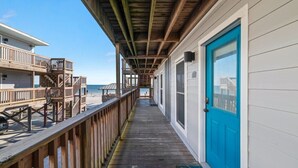 A wooden deck leading to a building with a blue door, a balcony, and a view of the beach.