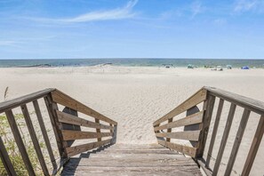Ein hölzerner Steg führt zu einem Sandstrand unter blauem Himmel.