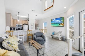 A modern kitchen with white cabinets, a dining area with wooden chairs, a gray sofa, and a TV mounted on the wall.