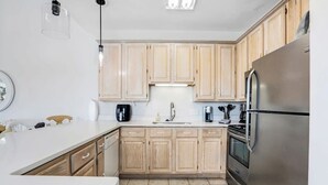 A modern kitchen with wooden cabinets, a stainless steel refrigerator, and a white countertop. - Camp David Side B- pool, beach view (Gulf Shores)