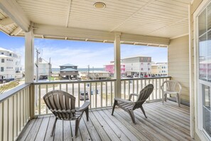 A covered porch with two Adirondack chairs and a table, overlooking a street with buildings and parked cars.