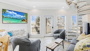 A modern living room with a large TV, comfortable seating, and a balcony with a view of the sea.