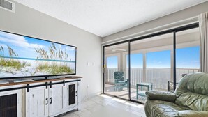 Un salón moderno con televisor de pantalla plana, un sofá de cuero y un ventanal corredero que da a un balcón con mobiliario exterior.