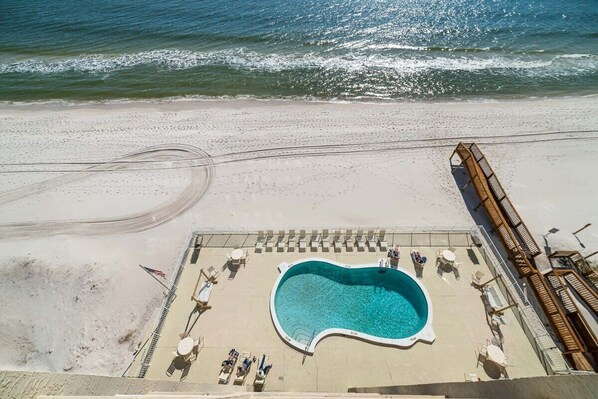 An aerial view of a beachside hotel with a swimming pool, lounge chairs, and a wooden deck overlooking the ocean.