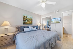 Une chambre à coucher avec un lit, deux lampes de chevet, un ventilateur de plafond, un tableau, une télévision et une porte.